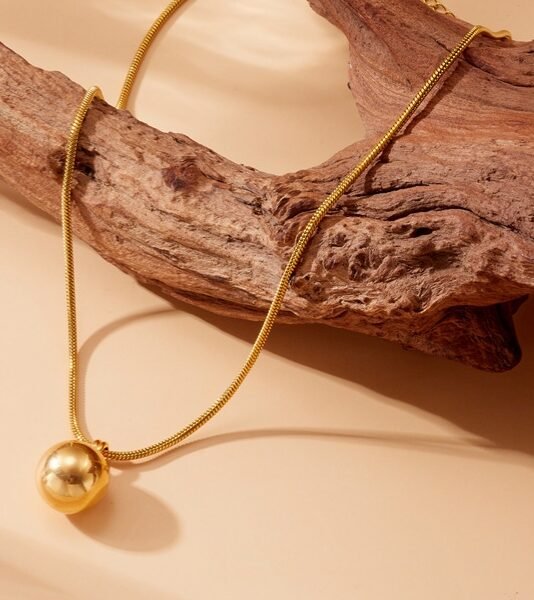 Minimalist gold sphere pendant necklace with a polished ball pendant on a sleek snake chain displayed on natural driftwood by https://vastorastore.com/